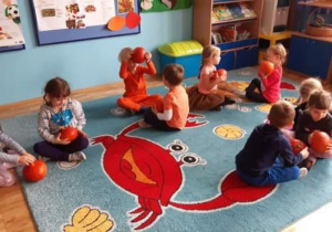 Dzieci siedzą w parach na dywanie i podają sobie dynie.