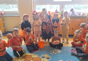 Dzieci stoją i siędzą w kręgu, są ubrane na pomarańczowo lub przebrane za dynie.