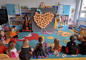 Dzieci siedzą naprzeciwko tablicy z balonami.