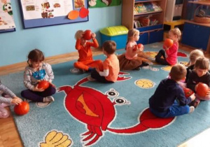 Dzieci siedzą w parach na dywanie i podają sobie dynie.