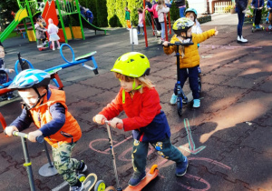 Dzieci jeżdżą na hulajnogach na przedszkolnym placu zabaw.