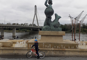 Dziecko stoi z rowerem przy pomniku Warszawskiej Syrenki.
