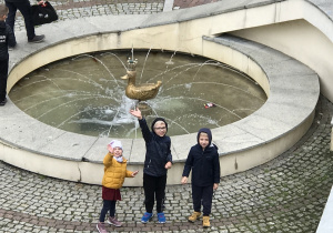 Troje dzieci stoi przy fontannie ze Złotą Kaczką.