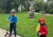 Dwoje dzieci stoi z hulajnogami przed pomnikiem przedstawiającym klucz wiolinowy.