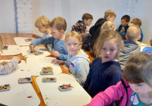 Dzieci siedzą przy długich stołach. Przed nimi leżą małe foremki z gotowymi kompozycjami kwiatowymi.