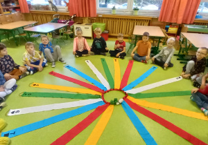 Dzieci siedzą w sali na podłodze w kole wokół wiatraka z kolorowych wstążek.