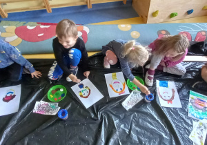 Dzieci siedzą na podłodze i malują farbami portrety kolegów i koleżanek.