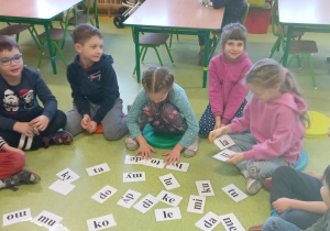 Dzieci na podłodze siedzą wokół kartek z sylabami.