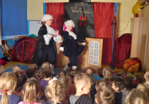 Teatrzyk. Dzieci oglądają dwie aktorki w białych perukach ubrane na czarno.