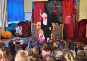 Teatrzyk. Dzieci oglądają aktorkę trzymającą dwie duże kukiełki w złotym i niebieskim ubraniu.