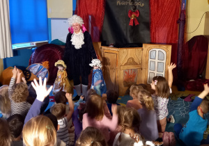 Teatrzyk. Dzieci oglądają aktorkę trzymającą dużą kukiełkę w różowym ubraniu.