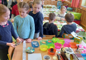 Dzieci stoją obok stolika i wybierają materiały plastyczne potrzebne do wykonanie sylwetki babci i dziadka.