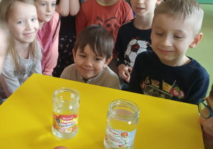 Dzieci siedzą wokół żółtego stołu i obserwują doświadczenie z jajkiem.