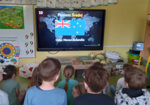 Dzieci siedzą przed dużym ekranem z flaga Nowej Zelandii.