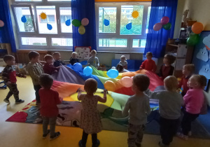 Dzieci stoją w kole z kolorową chustą i odbijają baloniki.