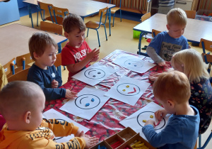 Dzieci siedzą przy stole i wyklejają plasteliną szablon uśmiechniętej emotikonki.