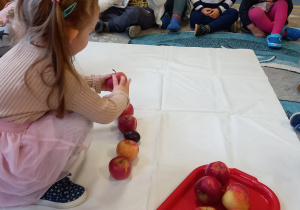 Dziewczynka układa owoce, pozostałe dzieci siedzą w kole.