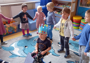 Urodziny. Dzieci stoją w kole, w środku siedzi chłopiec w niebieskiej koronie na głowie.
