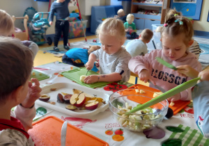 Dzieci siedzą przy stole i kroją owoce na sałatkę owocową.