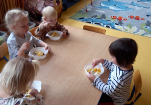 Dzieci siedzą przy stole i jedzą sałatkę owocową.