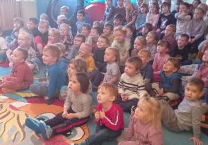 Duża grupa dzieci siedzi na dywanie i ogląda przedstawienie teatralne.