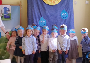 Pasowanie na Przedszkolaka. Dzieci z opaskami na głowie w kształcie kropelki wody stoją na tle dekoracji.
