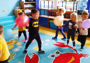 Dzieci bawiące się balonami na dywanie w sali przedszkolnej.