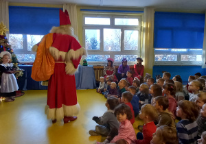 Dzieci siedzą na podłodze i patrzą na stojącego świętego Mikołaja.