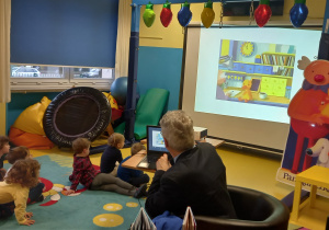 Spotkanie autorskie. Dzieci siedzą na dywanie o oglądają na dużym ekranie ilustracje do książek pana Widłaka.