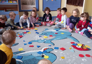 Dzieci siedzą wokół kolorowych figur geometrycznych rozłożonych na dywanie.