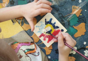 Dziewczynka maluje drewniany kafelek farbami.