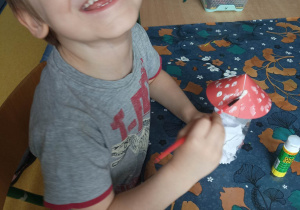 Chłopiec uśmiecha się malując farbą kapelusz muchomora.