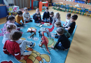 Dzieci siedzą w kole z figurkami dinozuarów.