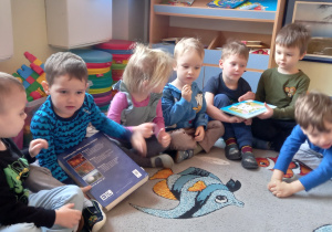Dzieci siedzą na dywanie i podają sobie książki