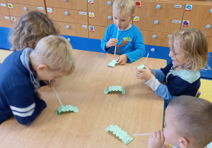 Dzieci siedzą przy stole i dmuchają przez słomki na gąsienice z papieru.