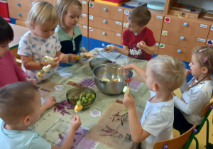Dzieci siedzą przy stole i robią szaszłyki z owoców.
