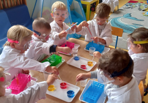 Zajęcia badawcze. Dzieci siedzą przy stole w białych fartuchach i nabierają pipetami kolorową wodę.
