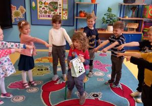 Dzieci stoją w kole i pokazują rękami na chłopca, który siedzi w środku koła z torebką prezentową z wzorem w pandy.