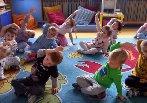 Dzieci siedzą na dywanie i wykonują skłon w bok z ręką uniesioną do góry.