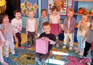 Dzieci stoją w kole, a w środku koła siedzi chłopiec z różowa torebką prezentową.