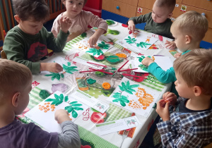 Dzieci siedzą przy stole i malują jarzębinę stemplując kartkę małą kuleczka umoczoną w farbie.