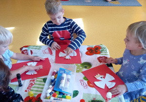 Dzieci siedzą przy stoliku i przyklejają orła w koronie zrobionego z papierowego talerzyka.