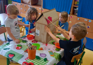 Dzieci siedzą przy stole i malują farbami domek z kartonu.