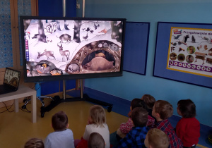 Dzieci oglądają na dużym ekranie film rysunkowy o przygotowaniach zwierząt do zimy.