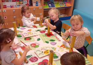 Dzieci siedzą przy stoliku i malują tekturowe rurki złotą farbą.
