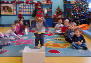 Dzieci siedzą na dywanie, a chłopiec w kolorowej czapce niesie obrazek do pudełka.