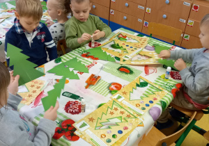 Dzieci siedzą przy stole i ozdabiają zielone papierowe choinki.