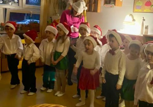 Dzieci w czapkach Mikołaja razem z nauczycielką stoją w sali, w której jest nastrojowe ciemne światło.