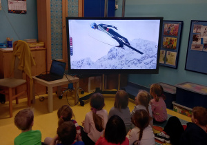Dzieci oglądają pokaz sportów zimowych na tablicy multimedialnej - skoki narciarskie.