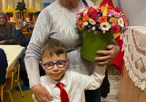Dzień Babci i Dziadka. Babcia stoi z wnuczkiem i trzyma kwiaty.